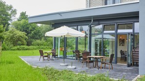 An outdoor patio with tables and chairs, a white umbrella, and a glass door entrance to a building. - VR-Serviced Apartments Obergeis (Neuenstein)