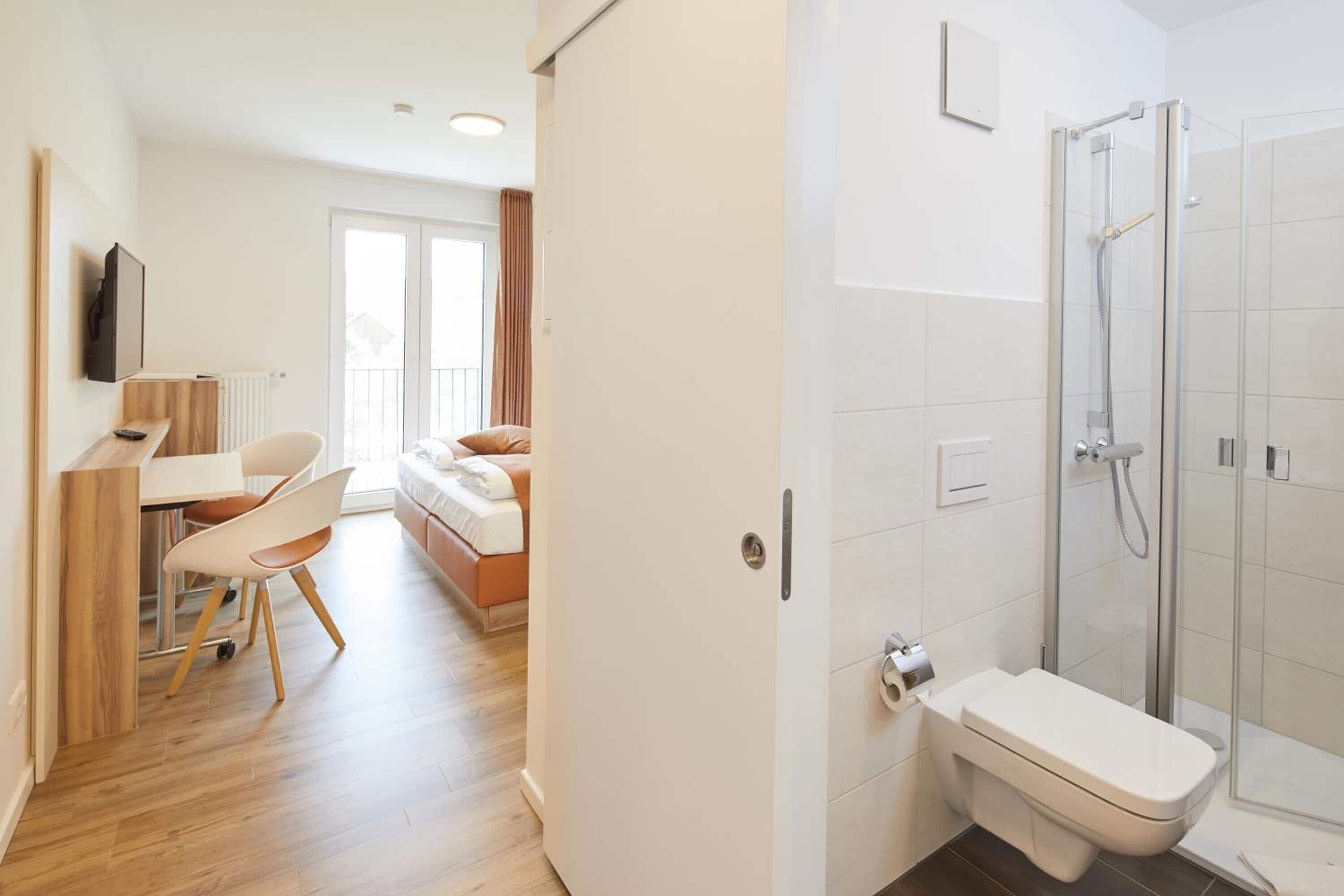 Una camera d'albergo moderna con pavimento in legno, una scrivania con sedia, un letto con testiera bianca e cabina doccia in vetro.