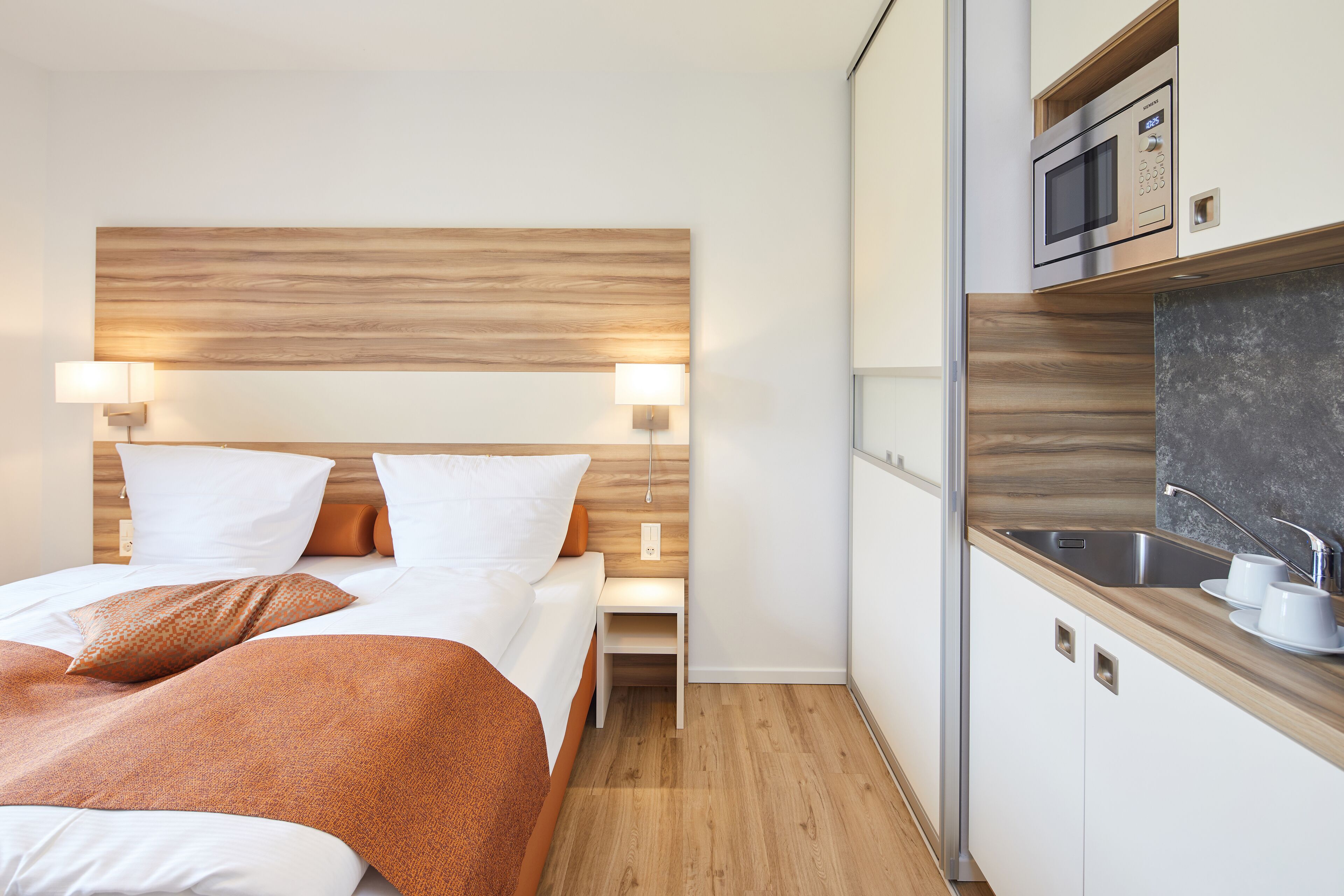 Una habitación de hotel con cabecera de madera, cama con ropa blanca, mesita de noche con lámpara y una kitchenette con microondas y lavamanos.