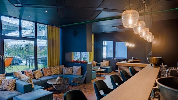 Un espace bar moderne doté d’un long comptoir en bois, de plusieurs options d’assises, notamment un canapé et des fauteuils, et d’une grande fenêtre offrant une vue sur l’extérieur.