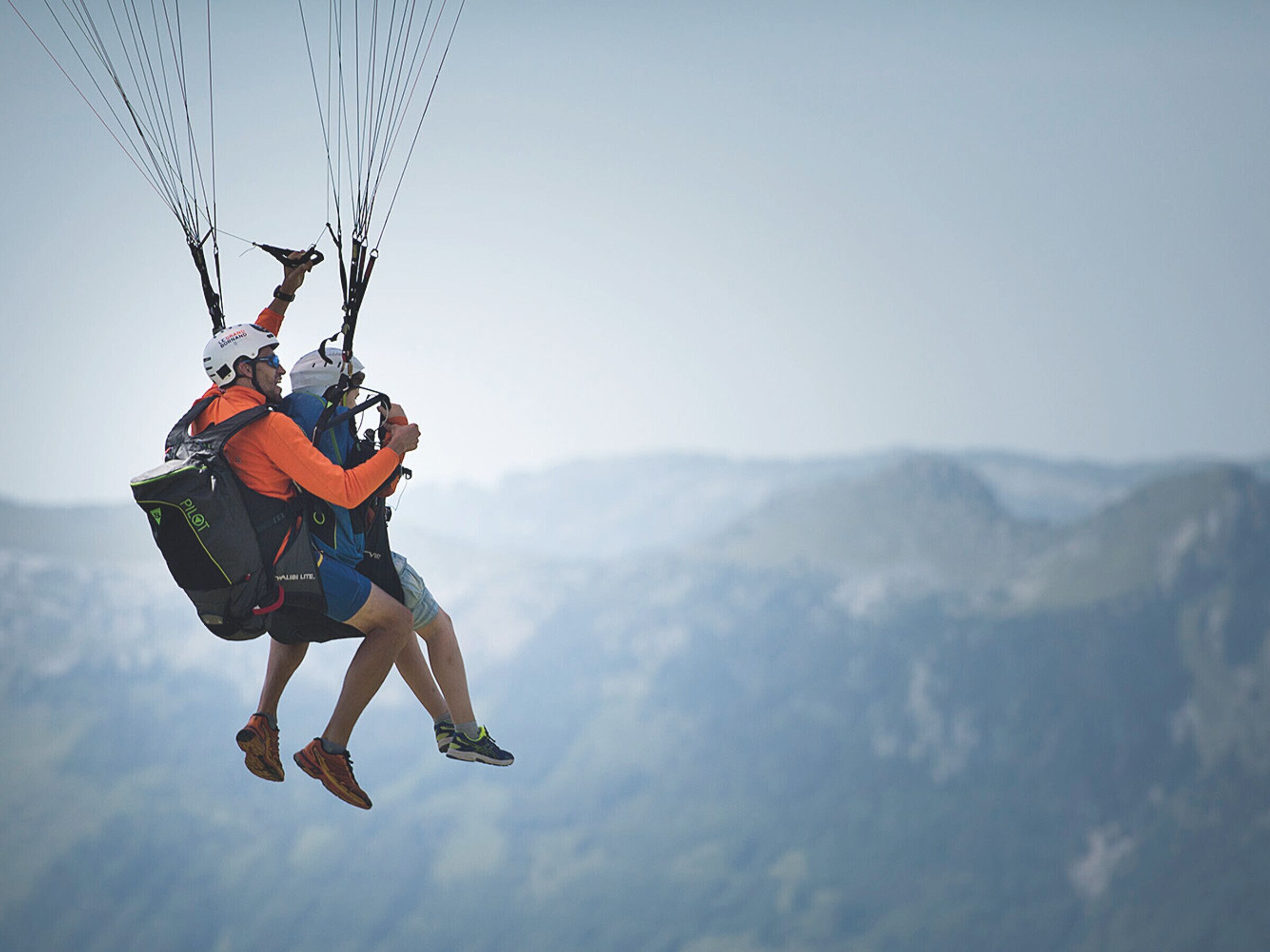 Two people paragliding with a mountainous landscape in the background.