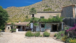 A stone building with a tiled roof, a small porch, and a garden with various plants and flowers. - BOQUER PETIT ( PIEDRA ). Surrounded by nature, but close to Pto. Pollensa (Puerto Pollensa)