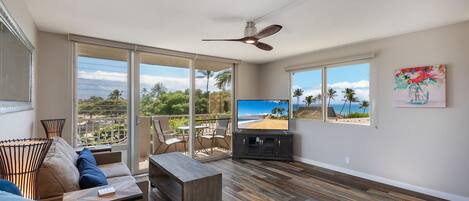 A spacious living area with a large TV, wooden flooring, and a balcony with a view of the beach.