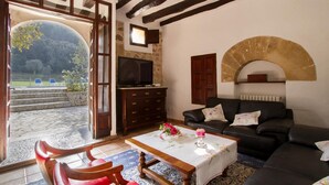 A living room with a stone fireplace, a black leather sofa, a wooden table with a vase of flowers, and a television on a wooden cabinet. - PLANA MORRO.  Characterful finca in beautiful countryside near Pollensa (Pollensa)