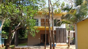 A modern two-story building with a yellow facade, a covered entrance, and a sandy courtyard. - Nature Breeze Resort (Vanur)