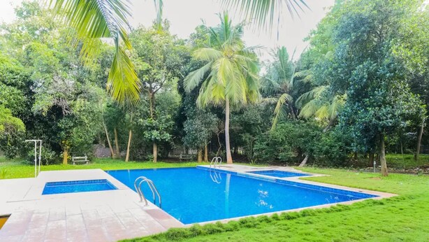 A rectangular swimming pool with a tiled deck, surrounded by lush greenery and palm trees. - Nature Breeze Resort (Vanur)