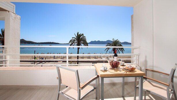 Un balcón con una mesa para dos, con vista a una playa con palmeras y un cielo azul despejado.