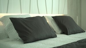 Una cama con cubrecolchón acolchado y dos almohadas, una oscura y otra clara, en una habitación con una pared lisa y una ventana con cortinas.