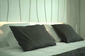 A bed with a quilted cover and two pillows, one dark and one light, in a room with a plain wall and a window with curtains.