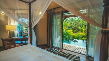 A bedroom with a bed, a desk, and a view of a lush garden and pool.