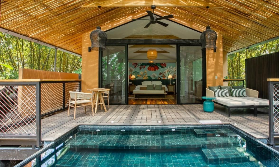 A poolside area with a wooden deck, a glass-enclosed bedroom, and a seating area with a sofa and chairs.