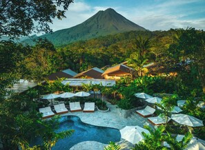 A resort with a swimming pool, lounge chairs, umbrellas, and a mountain in the background. - Nayara Gardens (La Fortuna)