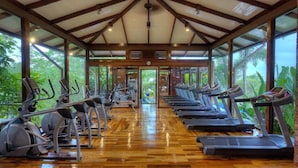 A gym with treadmills and elliptical machines, wooden flooring, and large windows with a view of greenery. - Nayara Gardens (La Fortuna)