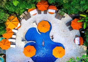 An aerial view of a circular pool with orange umbrellas and lounge chairs around it, surrounded by greenery.