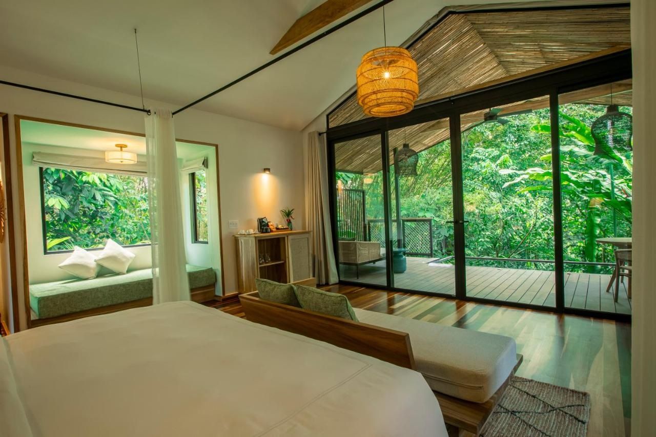 Une chambre moderne avec un grand lit, un coin salon avec des coussins, une petite table et une vue sur une végétation luxuriante grâce à de grandes fenêtres.