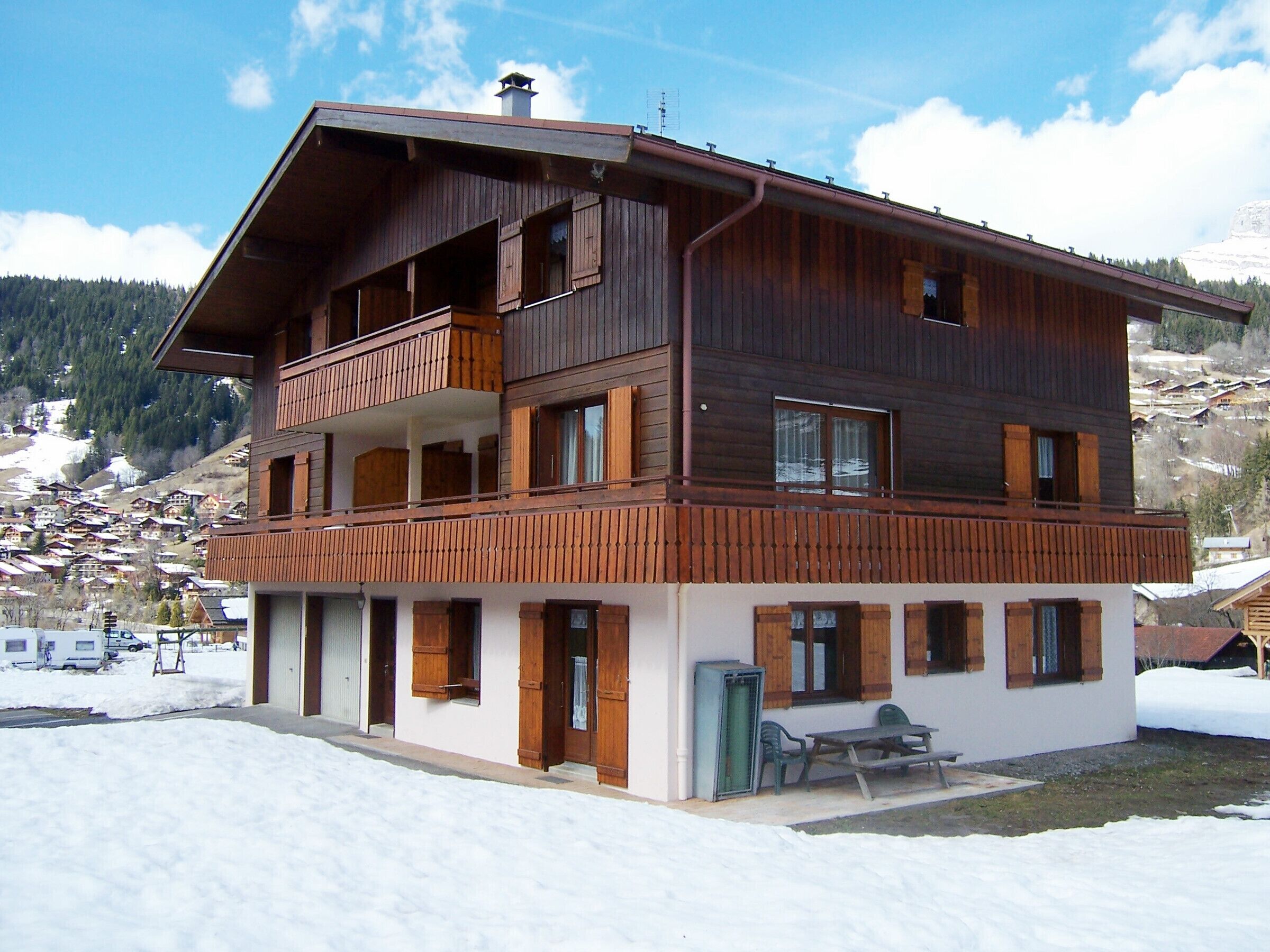 Eine zweistöckige Holzhütte mit einem Balkon im oberen Stockwerk, einer überdachten Außenbereich-Sitzgelegenheit und Blick auf eine verschneite Berglandschaft.