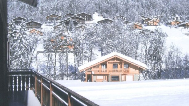 Une cabane en bois dotée d’un balcon, entourée d’arbres couverts de neige et d’autres cabanes situées sur une colline.