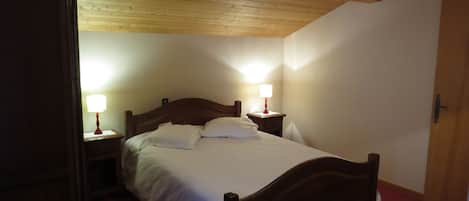 Quarto com teto de madeira, cama com lençóis brancos, duas luminárias de cabeceira e uma porta.