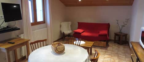 Un salon chaleureux avec une table ronde, des chaises en bois, un canapé rouge et un téléviseur posé sur un meuble.