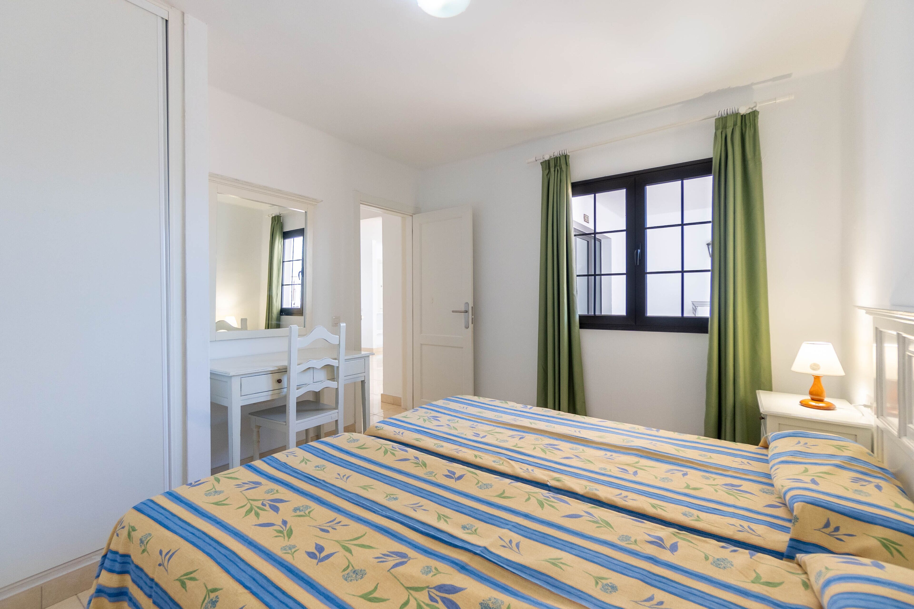 Quarto com cama, escrivaninha, cadeira, janela com cortinas e abajur.