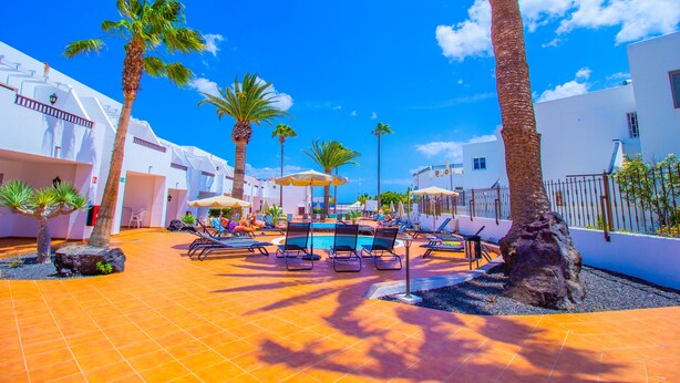 A poolside area with lounge chairs, palm trees, and a clear blue sky. - Apartamentos Flamingo (Tías)
