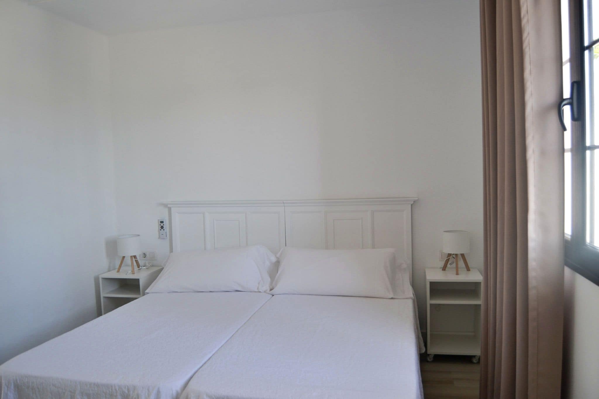 A double bed with white bedding in a room with white walls and a white headboard. There are two bedside tables with lamps.