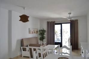 Una habitación con un sofá blanco, una mesa de comedor con sillas, un ventilador de techo, una planta y una ventana con cortinas.