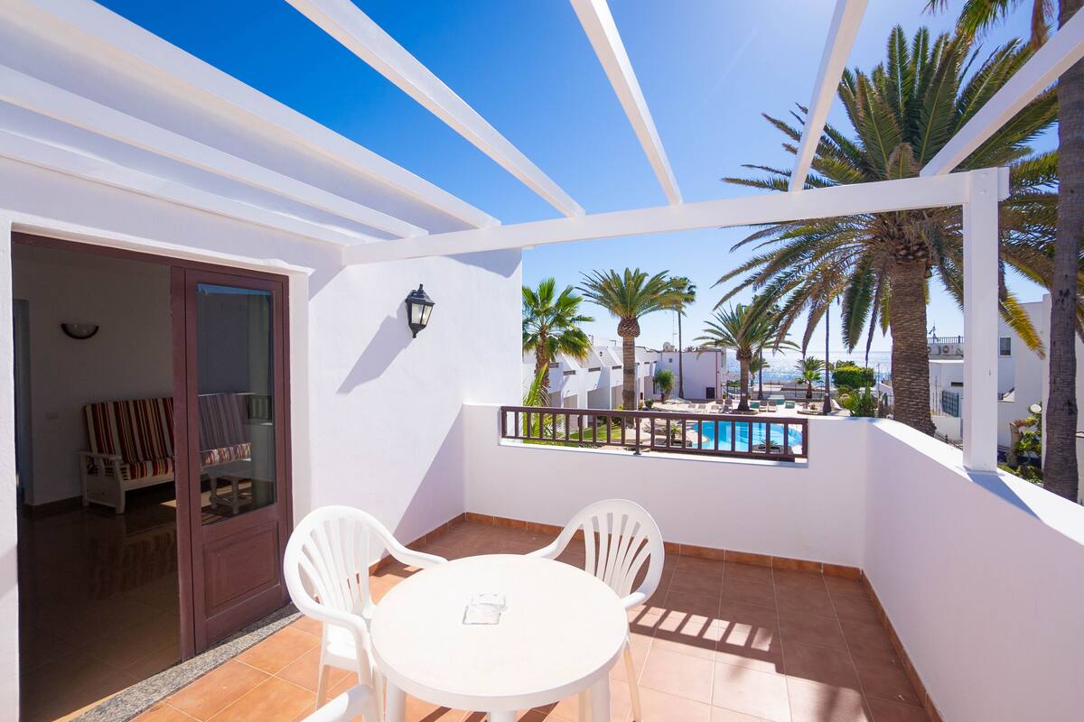 Un balcón con sillas y una mesa blancos, con vista a una piscina y palmeras.