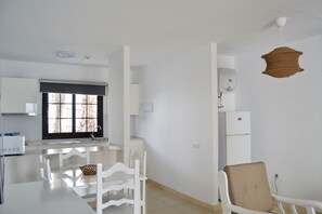 A modern kitchen with white cabinetry, a dining table with chairs, a window with a view, and a hanging light fixture. - Apartamentos Flamingo (Tías)