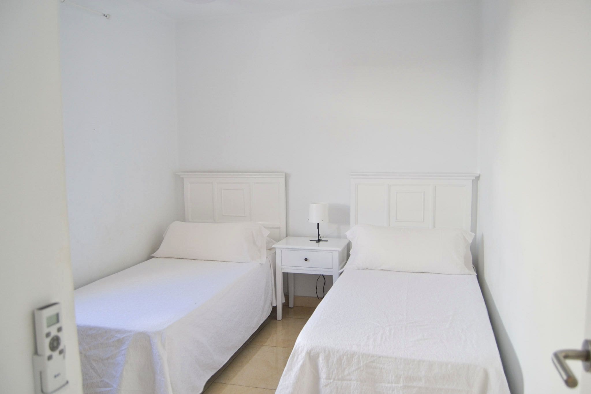 Una habitación con dos camas individuales, una mesita de noche con lámpara y un panel de control en la pared.