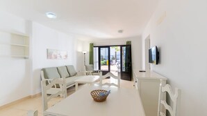 A modern living room with a white sofa, a dining table with a basket, and a TV mounted on the wall. - Apartamentos Flamingo (Tías)