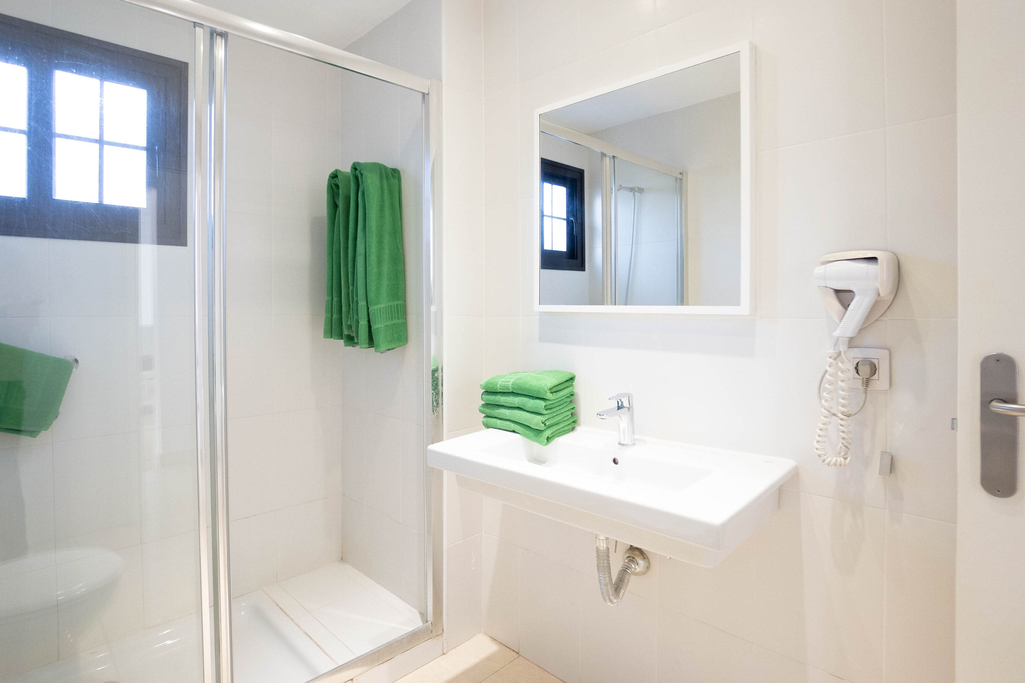 Une salle de bain avec une porte de douche en verre, un lavabo blanc, un miroir et un sèche-cheveux fixé au mur.