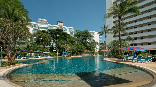 Una piscina amplia y cristalina con sillas de descanso y sombrillas, rodeada de palmeras y edificios modernos.