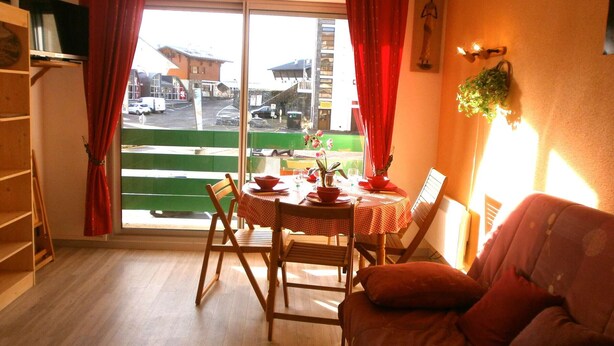 Ein Zimmer mit Esstisch für zwei Personen, einer Couch, einem Fenster mit roten Vorhängen und Blick auf eine Straße.