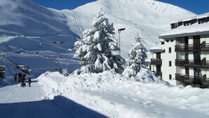 Ein mit Schnee bedeckter Berg mit einem Skigebiet im Hintergrund, ein Gebäude mit Balkonen und ein schneebedeckter Baum.