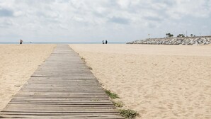 Una pasarela de madera que lleva a una playa arenosa con un litoral rocoso y palmeras a lo lejos.