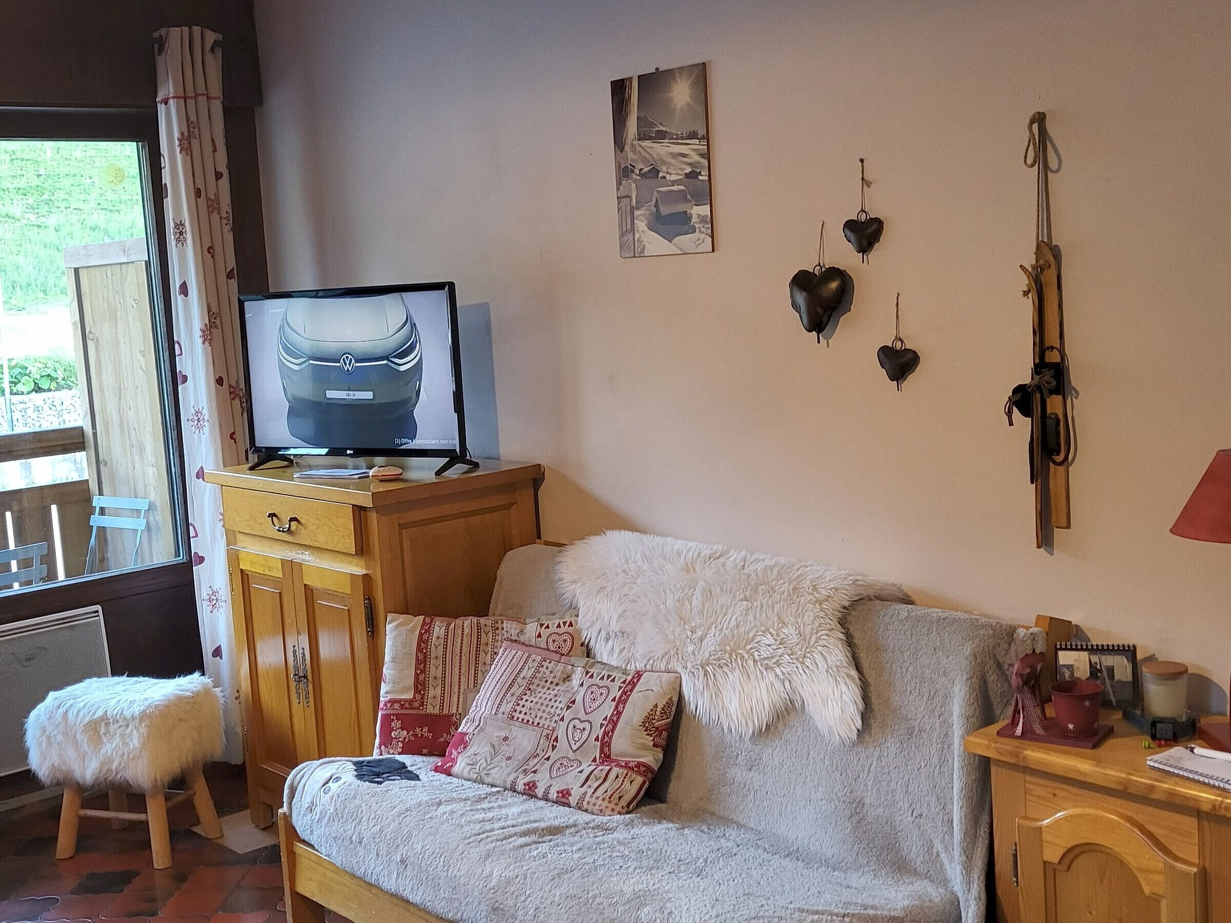 Ein gemütliches Wohnzimmer mit einer Couch, einem Fernseher auf einem Holzboden, einem kleinen Hocker und einem Fenster mit Vorhängen.