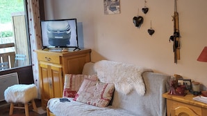 Una sala de estar acogedora con un sofá, un televisor sobre un mueble de madera, un taburete pequeño y una ventana con cortinas.
