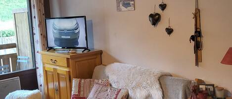 Ein gemütliches Wohnzimmer mit einer Couch, einem Fernseher auf einem Holzboden, einem kleinen Hocker und einem Fenster mit Vorhängen.