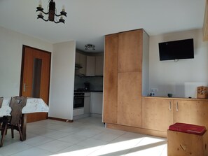 Ein Zimmer mit Esstisch, Holzschränken, einer Küchenzeile und einem an der Wand montierten Fernseher.