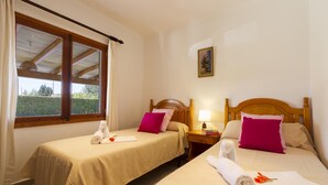 Ein Hotelzimmer mit zwei Betten, jeweils mit einem Holz-Kopfteil, beiger Bettwäsche und einem rosafarbenen Kissen. Es gibt ein Fenster mit Vorhängen, einen Nachttisch mit Lampe sowie ein Bild an der Wand.