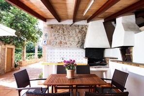 An outdoor patio with a stone wall, a wooden table, wicker chairs, and a potted plant. - PUNXA. Lovely stone-clad villa with large gardens surrounded by fruits and mountains (Pollensa)