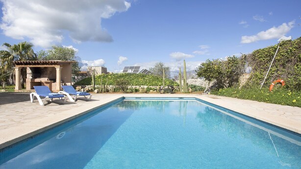 A rectangular swimming pool with clear blue water, surrounded by a tiled deck. There are two lounge chairs and a small covered seating area with a stone wall.