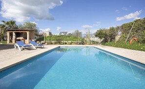 Et rektangulært swimmingpool med klart, blåt vand, omgivet af en flisbelagt terrasse. To liggestole og et lille overdækket siddeområde med en stenvæg.