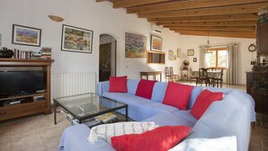 A living room with a sofa, coffee table, and television. There are books, paintings, and a dining area in the background.