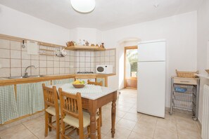 Een keuken met een betegelde spatwand, houten keukenkastjes, een spoelbak, een fornuis, een koelkast en een eettafel met stoelen.