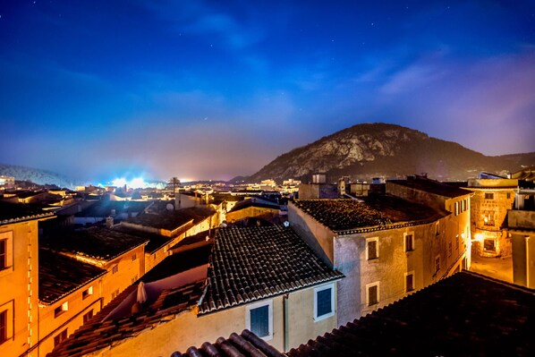 Vista noturna de uma cidade histórica com edifícios de telhados em terracota e uma colina imponente ao fundo.