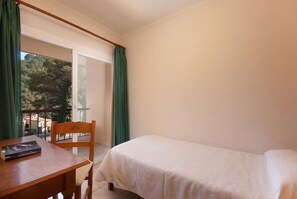 A room with a bed, a wooden chair, a table with a book, and a view of trees through a window.