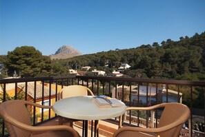 A balcony with wicker chairs and a table, overlooking a mountainous landscape and a village.