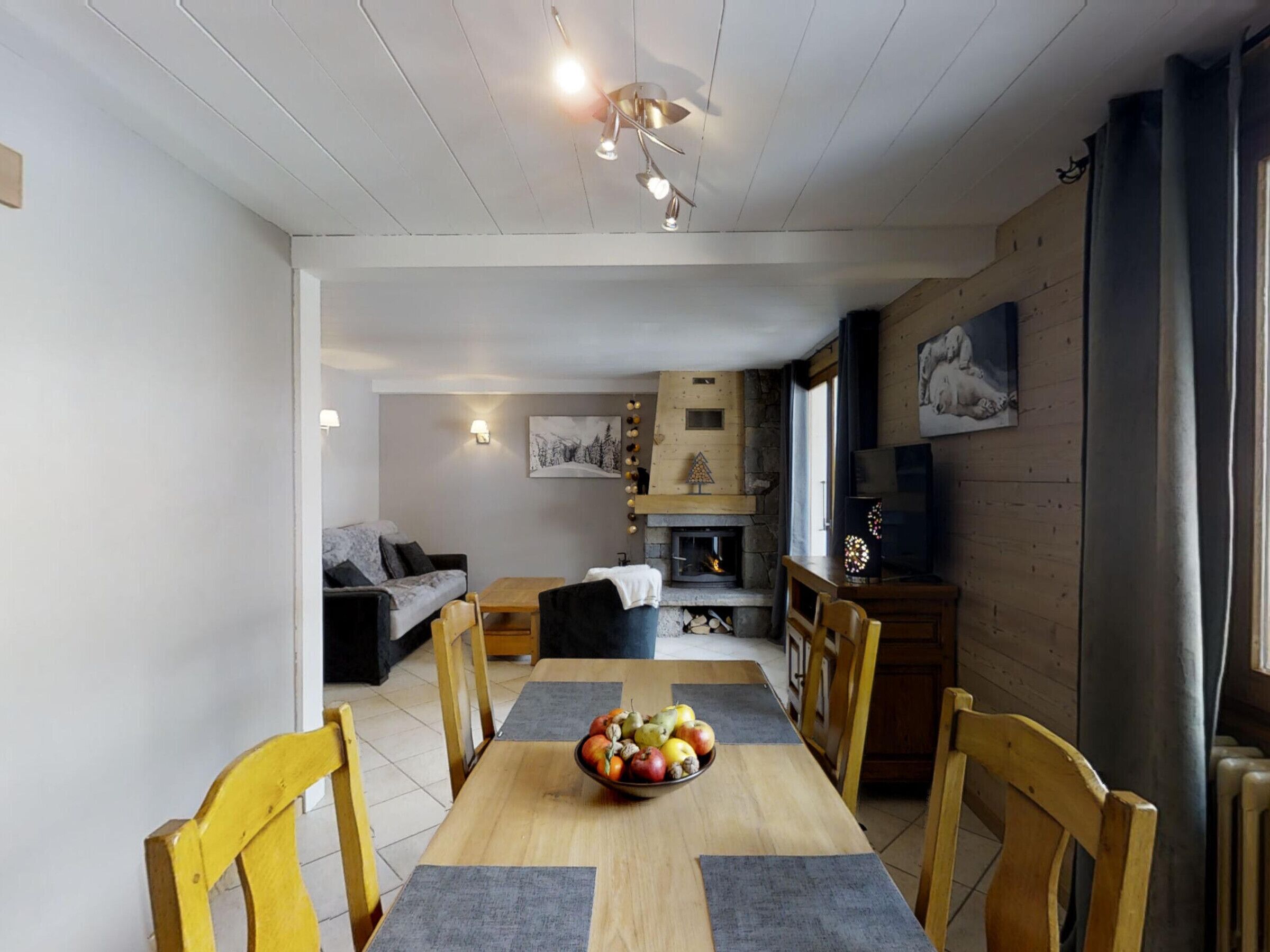 Un comedor con mesa de madera, sillas y un frutero. En el fondo, hay una chimenea, un televisor y un sofá.